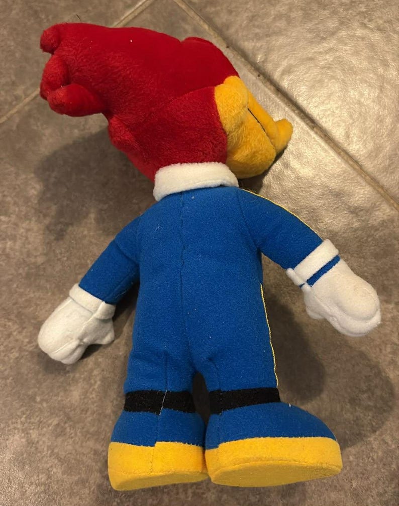 Puede incluir: Un peluche de un personaje de dibujos animados, un p&aacute;jaro carpintero, visto desde atr&aacute;s. El juguete tiene una cabeza roja, un pico amarillo y un mono azul con detalles amarillos. Guantes blancos y zapatos amarillos completan el atuendo. El juguete mide unos 25 cm de alto.