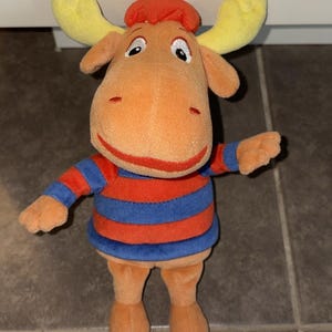 May include: A plush toy of a tan moose character with yellow antlers and a red tuft of hair on its head. The moose is wearing a shirt with horizontal red and blue stripes. It has large eyes, a wide red mouth, and small ears on the sides.