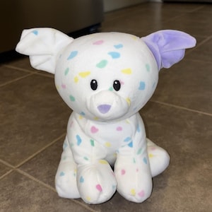 May include: A white plush dog toy with colourful confetti-like spots. The dog has black eyes, a purple nose, and one lavender ear. The toy is sitting on a tiled floor.