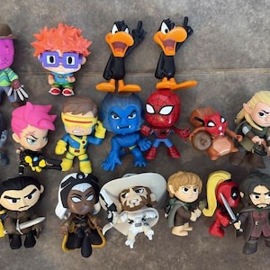 Peut inclure: Une collection de vingt figurines en vinyle stylisées représentant divers personnages de la culture populaire, incluant des super-héros, des figures fantastiques et des icônes de dessins animés, disposées en grille sur une surface carrelée.