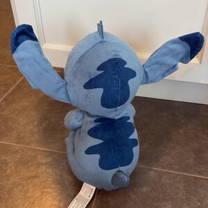 K&ouml;nnte beinhalten: Ein hellblaues Stitch-Pl&uuml;schtier mit dunkelblauen Markierungen und Akzenten. Das Spielzeug hat gro&szlig;e Ohren und einen kleinen Schwanz. Das Spielzeug befindet sich auf einem gefliesten Boden. Aus dem Film Lilo & Stitch.