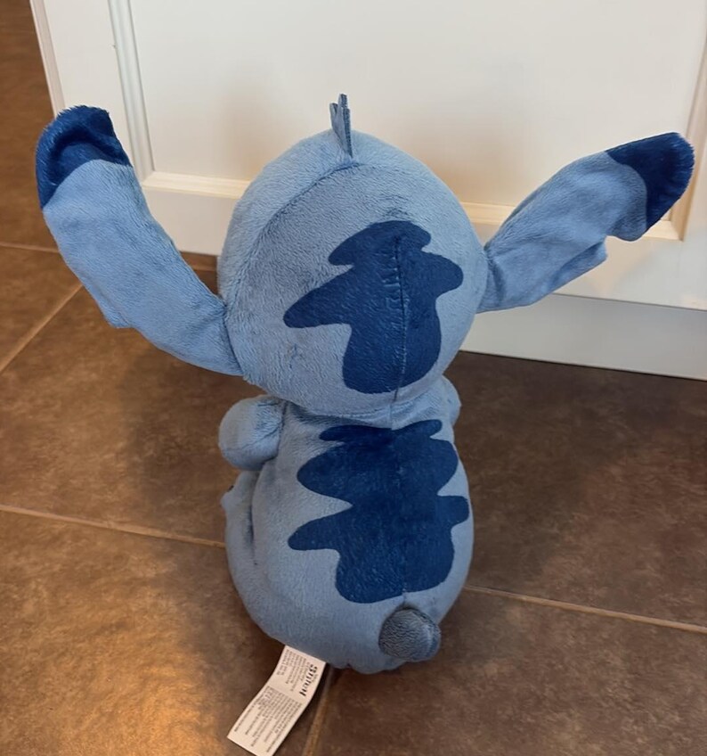 K&ouml;nnte beinhalten: Ein hellblaues Stitch-Pl&uuml;schtier mit dunkelblauen Markierungen und Akzenten. Das Spielzeug hat gro&szlig;e Ohren und einen kleinen Schwanz. Das Spielzeug befindet sich auf einem gefliesten Boden. Aus dem Film Lilo & Stitch.