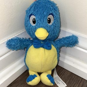 Peut inclure: Une peluche bleue avec une texture duveteuse, un ventre jaune, un bec jaune et un petit nœud papillon bleu. Le personnage a de grands yeux blancs avec des pupilles noires et des accents bleus. Il a des pieds jaunes avec des orteils bleus.