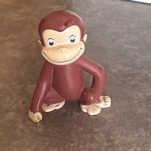 May include: A small, brown, moulded plastic figurine of a monkey with a light tan face, ears, hands, and feet. The monkey is depicted in a crouching position with a wide, gentle smile, large round eyes, and a smooth, matte finish throughout.