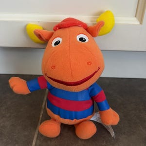 May include: An orange plush toy character with yellow antlers and a red cap. The character has large white eyes with black pupils and a wide red stitched smile. It wears a blue and red horizontally striped shirt with long orange sleeves.