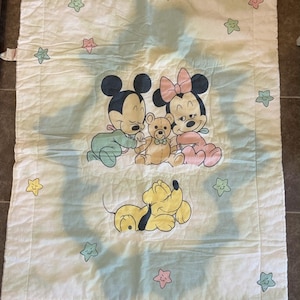Puede incluir: Una manta acolchada rectangular de color blanco con una ilustraci&oacute;n central de Mickey Mouse beb&eacute;, Minnie Mouse beb&eacute; y un osito de peluche, con Pluto debajo. El borde est&aacute; decorado con peque&ntilde;as estrellas de colores pastel rosa, amarillo y verde.