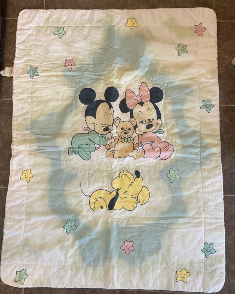 Puede incluir: Una manta acolchada rectangular de color blanco con una ilustraci&oacute;n central de Mickey Mouse beb&eacute;, Minnie Mouse beb&eacute; y un osito de peluche, con Pluto debajo. El borde est&aacute; decorado con peque&ntilde;as estrellas de colores pastel rosa, amarillo y verde.