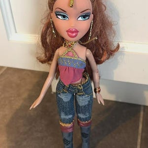 Puede incluir: Muñeca Bratz con cabello castaño y ojos azules, que lleva un top halter rosa y azul, vaqueros y botas grises. La muñeca tiene joyas doradas y una pieza de cabeza dorada. La muñeca está de pie sobre un suelo embaldosado.