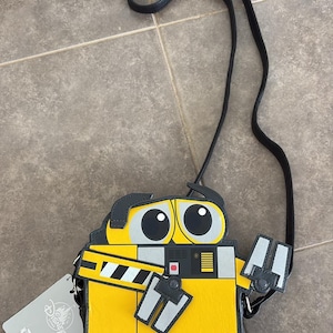 Può includere: Borsa a tracolla WALL-E gialla e nera con cinturino nero. La borsa presenta il design del viso e del corpo di WALL-E, con il nome "WALL-E" stampato sul davanti. Un'etichetta Disney è attaccata.