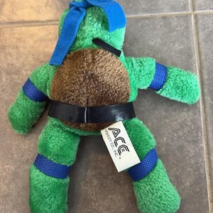 Peut inclure: Peluche verte repr&eacute;sentant une Tortue Ninja, avec une carapace marron et des d&eacute;tails bleus. Elle porte une ceinture noire et un masque bleu. Une &eacute;tiquette blanche avec "ACE NOVELTY CO., INC." est attach&eacute;e. Environ 20 cm de haut.