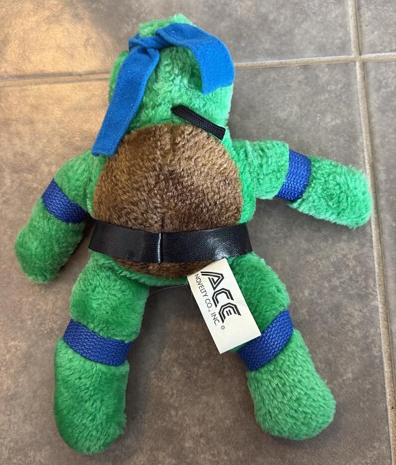 Peut inclure: Peluche verte repr&eacute;sentant une Tortue Ninja, avec une carapace marron et des d&eacute;tails bleus. Elle porte une ceinture noire et un masque bleu. Une &eacute;tiquette blanche avec "ACE NOVELTY CO., INC." est attach&eacute;e. Environ 20 cm de haut.