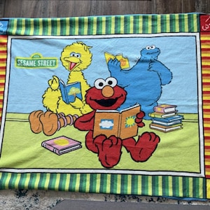 Op de afbeelding: Een Sesame Street-deken met Big Bird, Elmo en Cookie Monster die boeken lezen. De deken heeft een lichtblauwe achtergrond met het Sesame Street-logo en een groen, geel en rood gestreepte rand.
