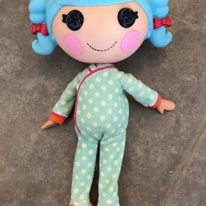 May include: A doll with bright blue hair styled in two pigtails with small red bows. The doll has large black button eyes, pink circular cheeks, and a stitched smile. It wears a light green long-sleeved onesie with white polka dots and pink slippers.