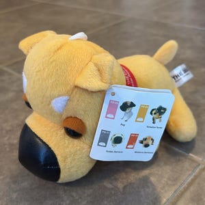 May include: A plush yellow dog toy with a black snout and a red collar. A tag shows images of different dog breeds, including a Pug, Yorkshire Terrier, Golden Retriever, and Miniature Dachshund. The toy is soft and cuddly.