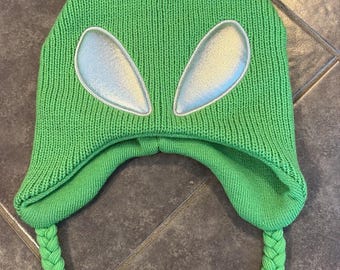 May include: A bright lime green knit beanie featuring two antennae with pom-poms on top, two large silver oval-shaped patches representing eyes, and two long braided earflaps with tassels at the ends, crafted from a textured yarn material.