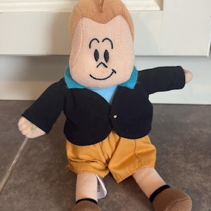 Könnte beinhalten: Eine Plüschpuppe mit hellbraunem Gesicht, gestickten Gesichtszügen, einer einzelnen Haarsträhne und einer weißen Mütze. Die Puppe trägt eine schwarze Jacke über einem hellblauen Hemd, senfgelbe Shorts und braune Schuhe mit schwarzem Rand.