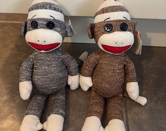 Peut inclure: Deux singes en peluche en forme de chaussettes. L'un est gris avec un chapeau rayé blanc et gris, un visage blanc et une bouche rouge. L'autre est marron avec un chapeau rayé blanc et marron, un visage blanc et une bouche rouge.