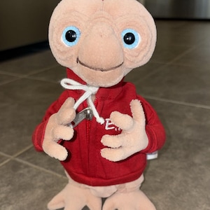 Op de afbeelding: Een knuffel van het personage E.T. met een lichtbruine huid, grote blauwe ogen en een gerimpeld hoofd. Het figuur draagt een rood vest met capuchon, een rits, witte trekkoorden en de letters E.T. in het wit op de borst geborduurd.
