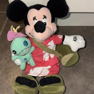 Puede incluir: Un peluche de Mickey Mouse con una camisa hawaiana roja con estampados florales blancos, una bandolera verde y zapatos verdes. Mickey sostiene un peque&ntilde;o peluche turquesa con un lazo rosa. El peluche tiene orejas negras y guantes blancos.