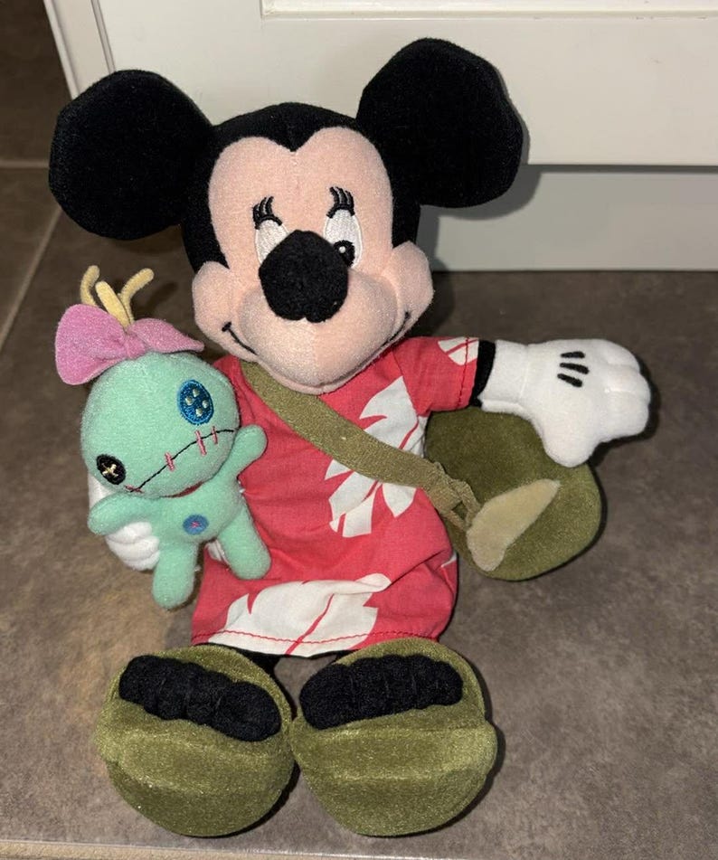 Puede incluir: Un peluche de Mickey Mouse con una camisa hawaiana roja con estampados florales blancos, una bandolera verde y zapatos verdes. Mickey sostiene un peque&ntilde;o peluche turquesa con un lazo rosa. El peluche tiene orejas negras y guantes blancos.