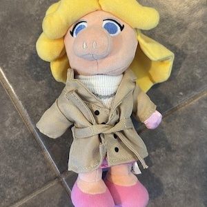 Könnte beinhalten: Ein Plüschtier von Miss Piggy aus den Muppets. Das Spielzeug hat gelbes Haar, blaue Augen und eine rosa Schnauze. Sie trägt einen beigefarbenen Trenchcoat, einen weißen Pullover und rosa Schuhe. Ein Sammlerstück.