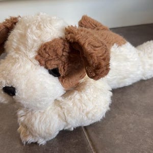 Può includere: Un cane di peluche con morbida pelliccia color crema e macchie marroni sulle orecchie e intorno agli occhi. Il giocattolo ha un naso nero, occhi scuri ed è raffigurato in posizione sdraiata con le zampe anteriori ripiegate sotto il corpo.