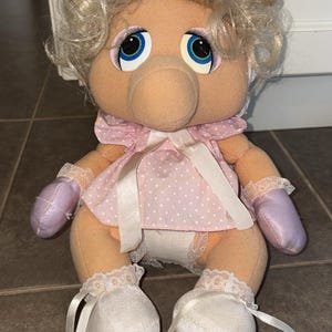 May include: A plush toy of a cartoon pig character with blonde curly hair, blue eyes, and a pink dress with white polka dots. The doll has lavender gloves and white shoes with ribbons.