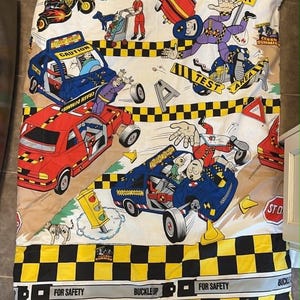 Puede incluir: Una sábana beige y blanca con un diseño de dibujos animados colorido. El diseño presenta coches, motocicletas y figuras en escenarios de pruebas de choque. La sábana tiene texto que dice "CAUTION", "TEST AREA", "FOR SAFETY" y "BUCKLEUP".