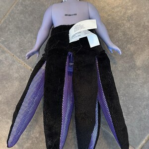 May include: A doll depicting Ursula from The Little Mermaid. The doll has a purple body, white hair, and a black skirt with purple and white accents. The doll's skirt is designed to resemble tentacles.