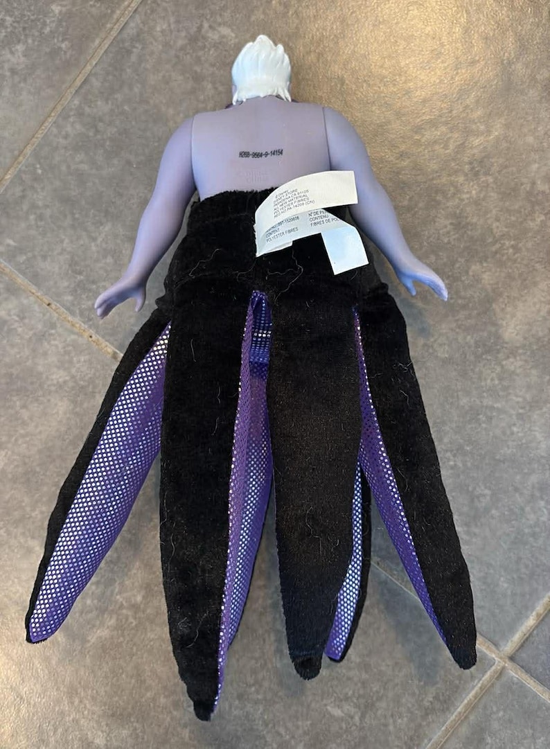 May include: A doll depicting Ursula from The Little Mermaid. The doll has a purple body, white hair, and a black skirt with purple and white accents. The doll's skirt is designed to resemble tentacles.
