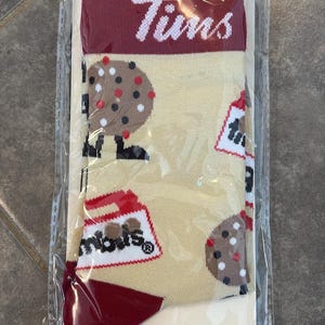 Può includere: Un paio di calzini beige con un motivo di biscotti con gocce di cioccolato e tazze di caffè, confezionati in plastica trasparente. La parte superiore dei calzini è bordeaux con la scritta "Tims" in bianco.