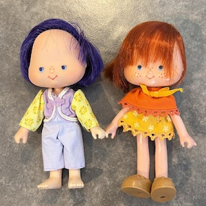 Puede incluir: Dos muñecas vintage con caras pintadas y cabello enraizado. Una muñeca tiene cabello morado, una chaqueta floral amarilla, un chaleco morado y pantalones azules. La otra muñeca tiene cabello rojo, una blusa naranja y una falda floral amarilla. Ambas muñecas se toman de la mano.