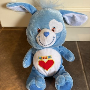 Puede incluir: Un peluche azul con forma de perro con una mancha blanca en la cabeza y un corazón rojo en el vientre. El perro tiene orejas caídas, una cara sonriente y un mechón de pelo azul. Tiene una nariz en forma de corazón y un emblema dorado y azul encima del corazón.