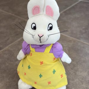 Könnte beinhalten: Ein Plüschhasen-Spielzeug mit weißem Fell, rosa Innenohren sowie schwarz gestickten Augen und Schnurrhaaren. Der Hase trägt ein violettes Kurzarmshirt unter einem gelben Latzkleid mit einem kleinen, sich wiederholenden, bunten Kreuzmuster.