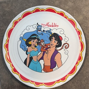 May include: Round white plate featuring characters from Disney's Aladdin. The plate has a red and yellow scalloped border. Aladdin, Jasmine, the Genie, and Abu are depicted in vibrant colours.