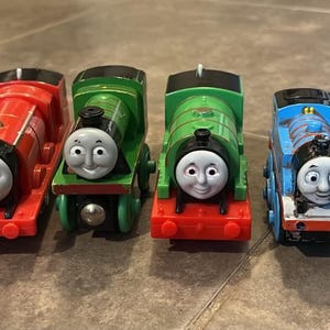 May include: A collection of five toy train engines and one red coal tender. The set includes a red engine with the number 5, a green engine, a second green engine, a blue engine, and a dark green engine, all featuring smiling faces.