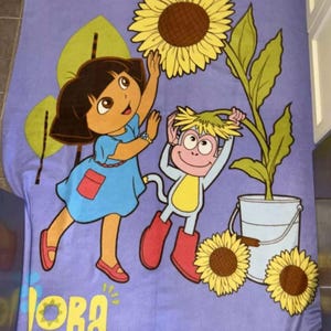 Puede incluir: Una manta de forro polar morada con Dora la Exploradora y Botas el Mono. Dora lleva un vestido azul y zapatos rojos. Botas lleva una camisa amarilla y botas rojas. Están de pie frente a un girasol.
