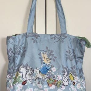 May include: A light blue tote bag featuring a printed pattern of Peter Rabbit characters, including a squirrel, a rabbit in a blue jacket, and a smaller rabbit, surrounded by white flowers, green leaves, a butterfly, and a ladybug.