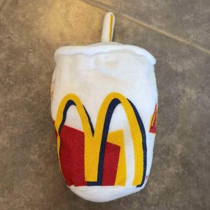 Könnte beinhalten: Weißes Plüschtier in Form eines McDonald's-Bechers mit gelbem Strohhalm. Der Becher trägt das McDonald's-Logo in Rot, Gelb und Blau.