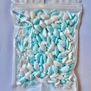 May include: A clear plastic bag filled with cloud-shaped sprinkles in shades of light blue and white. The sprinkles have a pearlescent sheen and are tightly packed within the sealed bag. The bag has a small hole at the top for hanging.