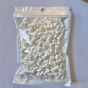 May include: A clear plastic bag filled with numerous white bone-shaped sprinkles. The sprinkles have a pearlescent sheen, giving them a slightly iridescent appearance. The bag is sealed at the top and is sitting on a white surface.