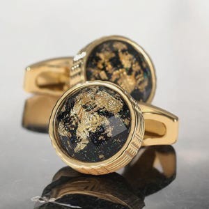 May include: A pair of gold-toned cufflinks with a round, black center embedded with gold leaf. The cufflinks have a textured, ridged design around the black center and a simple, curved bar for attachment.