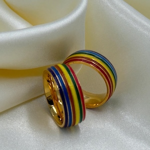 May include: Two gold-colored rings with a rainbow stripe design. The rings have a band of red, orange, yellow, green, blue, and purple stripes. The rings are set against a white satin background.