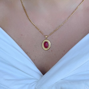 Könnte beinhalten: Goldfarbene Halskette mit einem roten, ovalen Anhänger. Der Anhänger ist in einen goldenen Rahmen gefasst und hängt an einer filigranen Kette. Die Halskette wird vor weißem Hintergrund gezeigt.