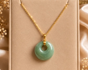 May include: A gold chain necklace with a circular, light green jade pendant. The pendant has a hole in the center. The necklace is displayed on a beige jewelry box, with a satin background and dried flowers.