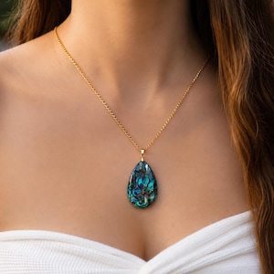 Peut inclure: Un pendentif en forme de goutte orné d'une incrustation de coquille d'ormeau irisée aux motifs tourbillonnants bleu profond, vert et noir, suspendu à une fine chaîne en métal doré reposant sur la clavicule.