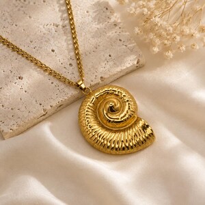 May include: A gold-toned necklace featuring a pendant shaped like a spiraled ammonite fossil. The pendant has a polished, reflective surface with detailed ridges and grooves, suspended from a matching gold-toned metal chain link necklace.