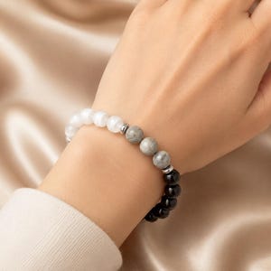 May include: A beaded bracelet featuring a sequence of smooth white, grey, and black spherical beads. The design includes small metallic silver-toned spacers placed between the different colored bead sections, creating a distinct contrast in the arrangement.