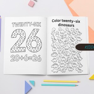 May include: An open children's activity book with the text "TWENTY-SIX" and "20+6=26" on one page, and "Color twenty-six dinosaurs" with dinosaur illustrations on the other. Colored pencils and geometric shapes are scattered around the book.
