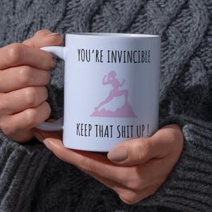 May include: White ceramic mug with the text "YOU'RE INVINCIBLE" and "KEEP THAT SHIT UP!" in black. A pink silhouette of a person running on a mountain is in the center. The mug is held by a person wearing a gray sweater.